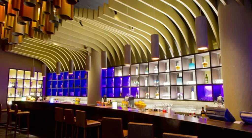 Dalgalı ahşap akustik tavan kaplaması olan bar/lounge iç mekanı, şişe rafları ve tabureler