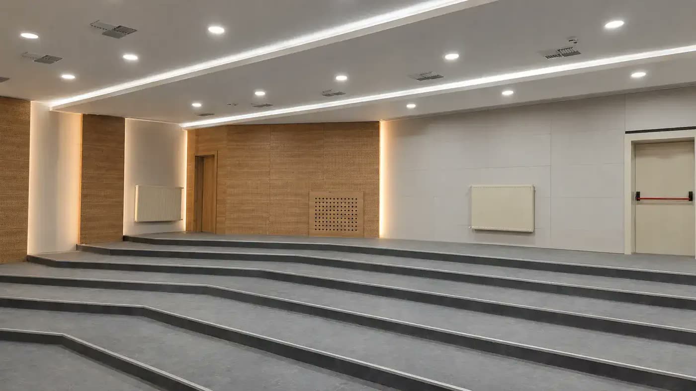 Basamaklı oturma düzenine sahip konferans salonu; duvarlarda dokulu/delikli ahşap desenli akustik panel şeritleri ve aralarında radyatörler görünüyor